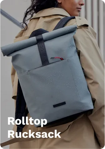 Gray rolltop rucksack with black straps and red accents, worn by a person, standing outdoors near a stone building.