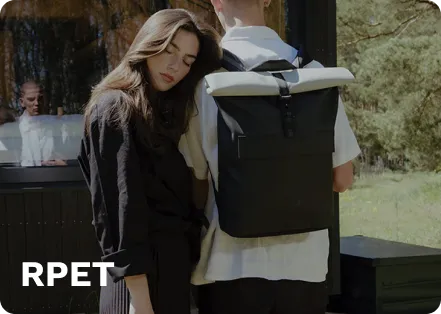 Black RPET roll-top backpack with gray top, worn by a person, standing outdoors near a wooden structure with greenery in the background.