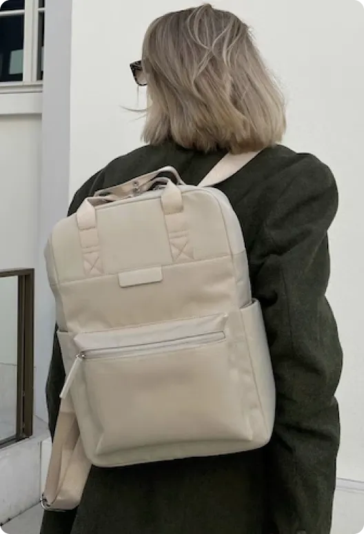 Beige backpack with top handles, zippered pocket, and adjustable straps, worn by a person, designed for everyday use.