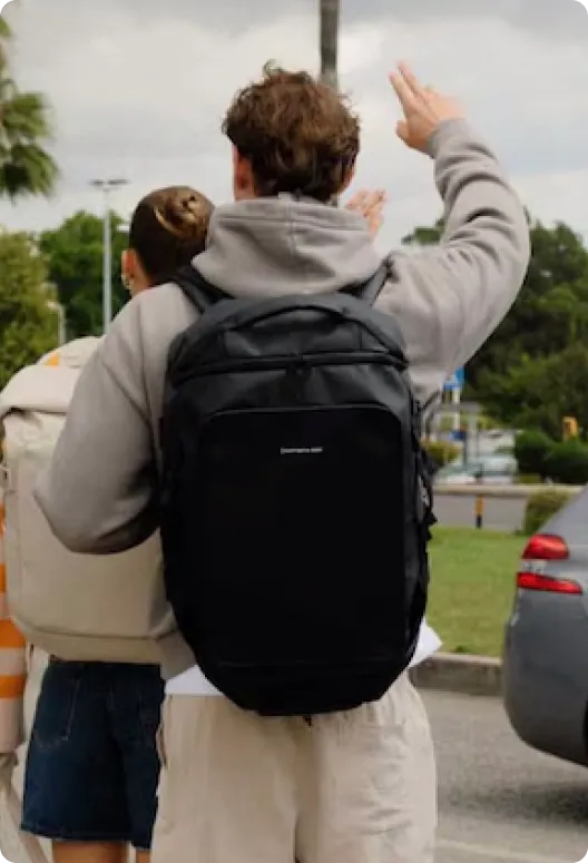 Black duffle backpack with a branded logo, worn by a person raising a hand, standing outdoors near a road with greenery and a car in the background.