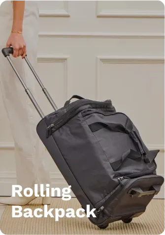 Black rolling backpack with wheels, telescopic handle, and zippered compartments, pulled by a person on a carpeted floor near a white door.