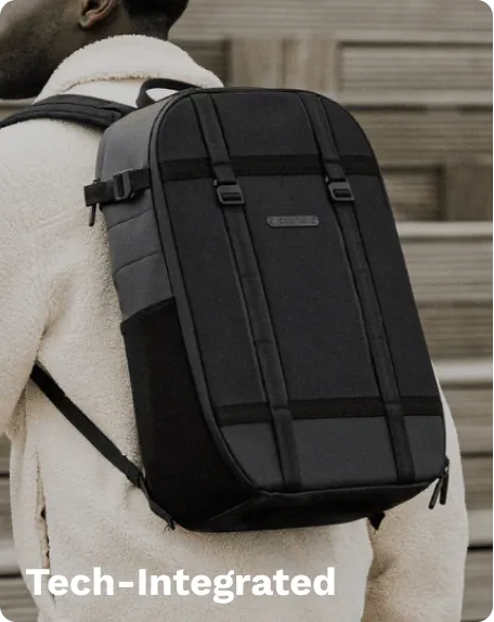 Black tech-integrated backpack with gray panels, multiple straps, and a branded logo, worn by a person, standing outdoors near a stone wall.