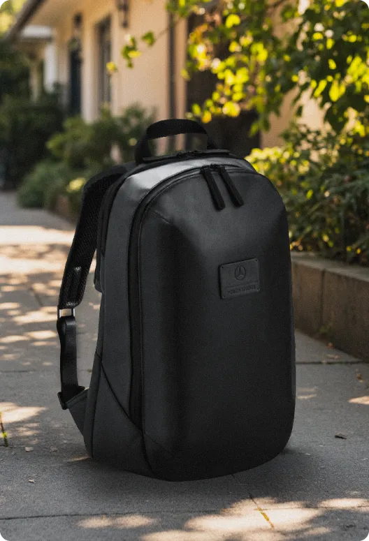 Sleek black backpack with top handles, dual zippered compartments, and adjustable straps, featuring a branded logo, on a sidewalk near green plants.