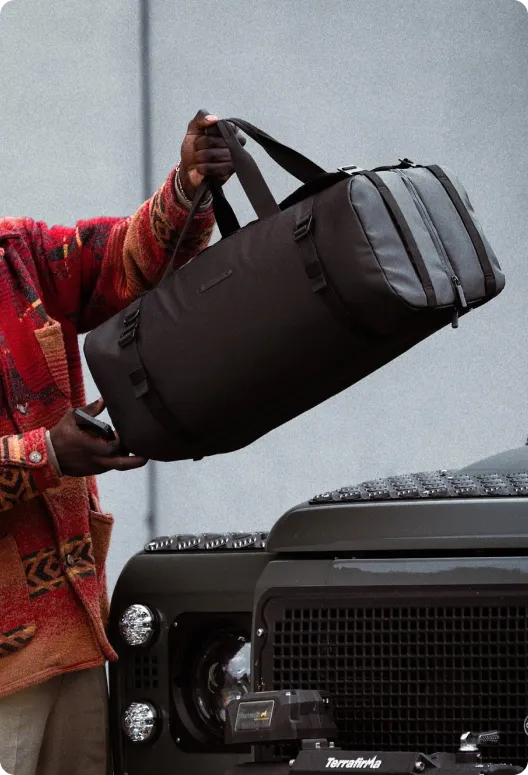 Black duffle bag with handles and zippers, held by a person, positioned in front of a black vehicle against a gray background.