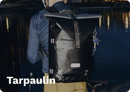 Black tarpaulin roll-top backpack with gray straps, worn by a person on a bicycle, near water at night with city lights reflecting.