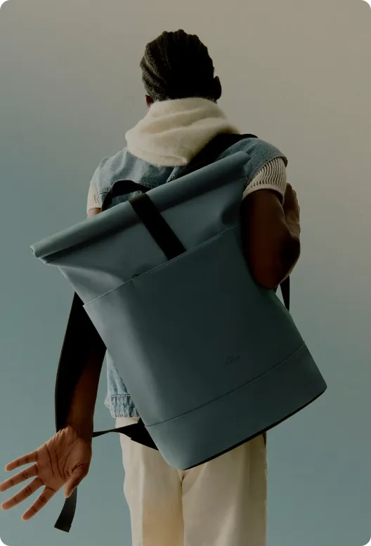 Blue roll-top backpack with black straps and handles, worn by a person in a white jacket and beige pants, against a light blue background.