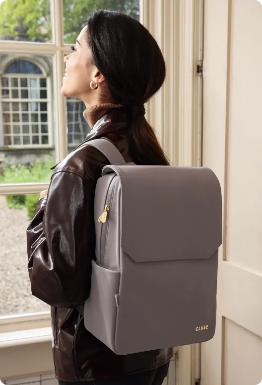 Gray leather backpack with gold zippers and a branded logo, worn by a woman, standing near a window with greenery outside.